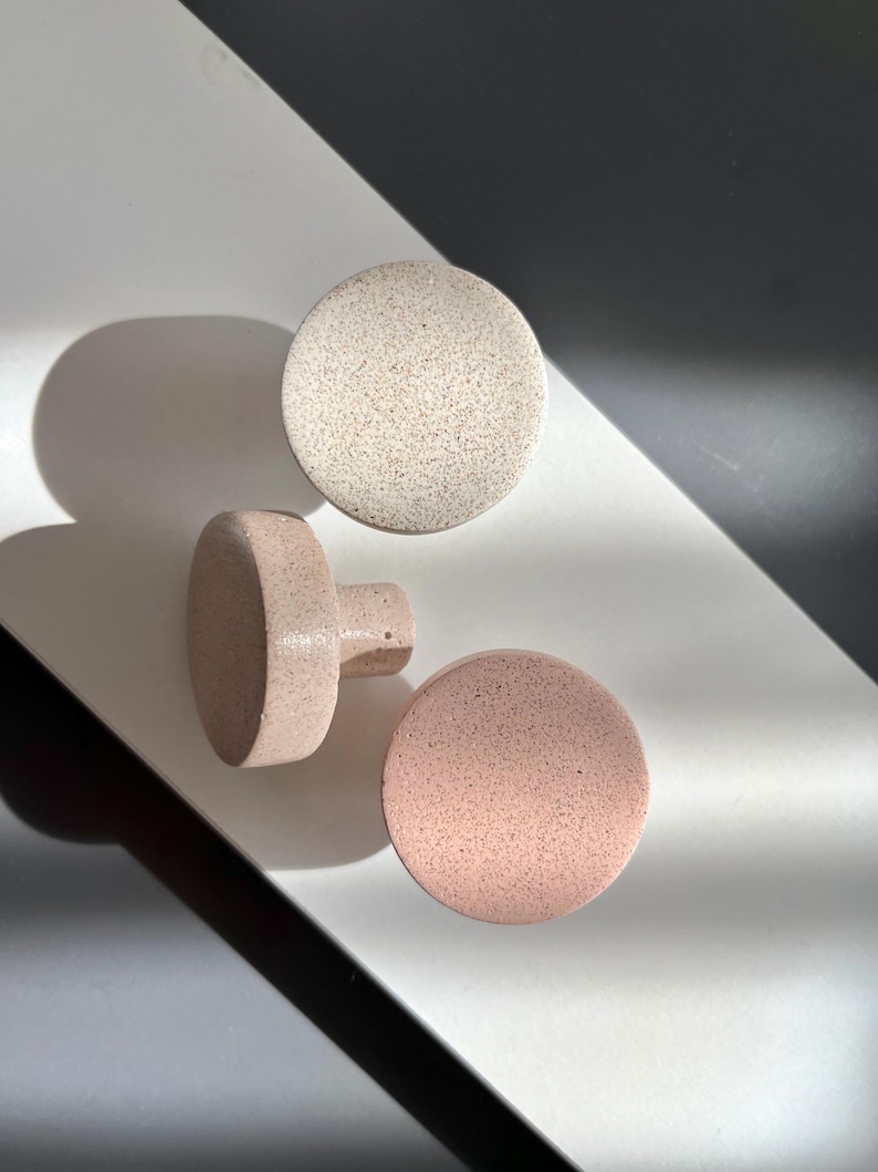 Puede incluir: Tres pomos de armario redondos y moteados en tonos neutros. Un pomo es de color beige claro y los otros dos son de color rosa p&aacute;lido. Un pomo rosa tiene un v&aacute;stago cil&iacute;ndrico. Los pomos est&aacute;n dispuestos sobre una superficie blanca y negra.