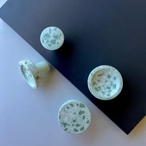 Könnte beinhalten: Vier hellgrüne Terrazzo-Schrankknöpfe und eine kleine Schale. Die Knöpfe haben eine runde Form mit einem gesprenkelten Muster aus Weiß, Grün und dunklerem Grün. Die Schale hat ein ähnliches gesprenkeltes Muster.
