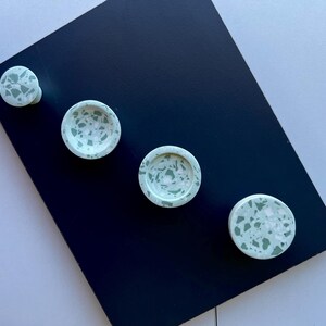 May include: A collection of four light green and white terrazzo-style decorative knobs and small dishes. The knobs and dishes are arranged on a dark blue surface, with a white background.