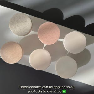 Puede incluir: Seis objetos redondos en tonos neutros, beige, rosa y blanco, dispuestos sobre una superficie blanca. La imagen incluye texto que indica que estos colores est&aacute;n disponibles para todos los productos. Los objetos est&aacute;n espaciados uniformemente y tienen un acabado mate.