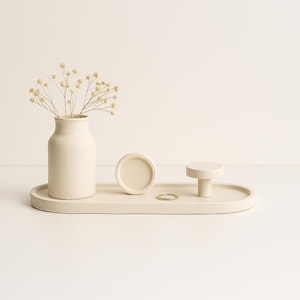 May include: A white oval tray with a white ceramic vase, a white round dish, and a white knob. The vase holds dried flowers. A gold ring is on the tray.