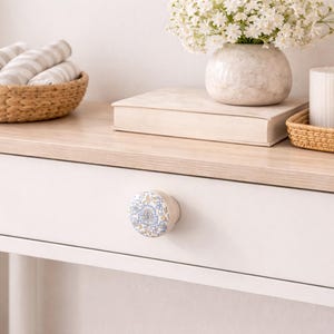 Op de afbeelding: Een wit geschilderde houten consoletafel met een licht houten blad. Een lade met een bloemvormige knop staat in het midden. Een geweven mand met opgerolde handdoeken, een vaas met witte bloemen, een kaars en een dienblad staan erop.