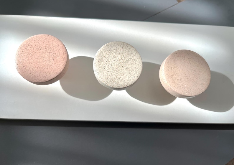 Puede incluir: Tres objetos redondos y texturizados en tonos rosa y beige est&aacute;n dispuestos sobre una superficie blanca. Los objetos proyectan sombras, lo que sugiere una fuente de luz desde arriba. Los art&iacute;culos parecen ser productos de ba&ntilde;o o belleza.