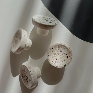 May include: Four terrazzo cabinet knobs. Each knob features a white base and a speckled pattern of black, pink, and beige. The knobs are round with a short, cylindrical stem. The knobs are designed for use on cabinets or drawers.
