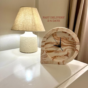 May include: A decorative clock with a cream and brown marbled face and black hands. The clock is on a white surface next to a cream-coloured lamp with a beige lampshade. The text "FAST DELIVERY 2-4 DAYS" is visible.