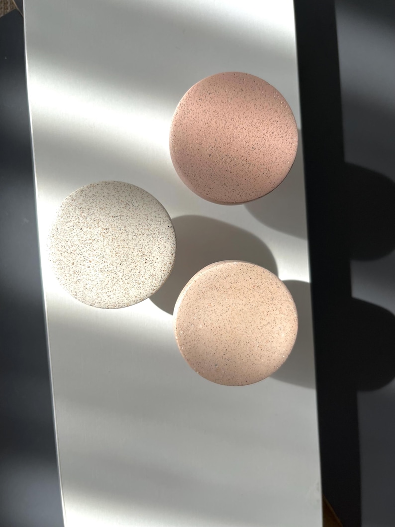 Puede incluir: Tres posavasos redondos y moteados en tonos blanco, rosa y beige. Los posavasos est&aacute;n dispuestos sobre una superficie clara, con sombras. El dise&ntilde;o moteado les da una apariencia texturizada, adecuados para la decoraci&oacute;n del hogar.