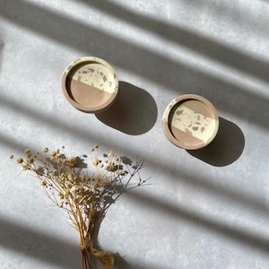 May include: Two small round concrete coasters with a white and beige speckled design. The coasters are on a light grey surface with a bouquet of dried white flowers.