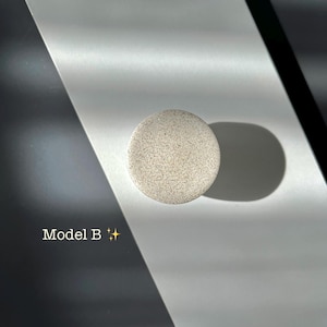 Puede incluir: Un objeto redondo, moteado, gris claro, etiquetado como "Modelo B" con un gr&aacute;fico de brillo. El objeto proyecta una sombra sobre una superficie blanca y gris. La imagen est&aacute; bien iluminada con sombras fuertes.