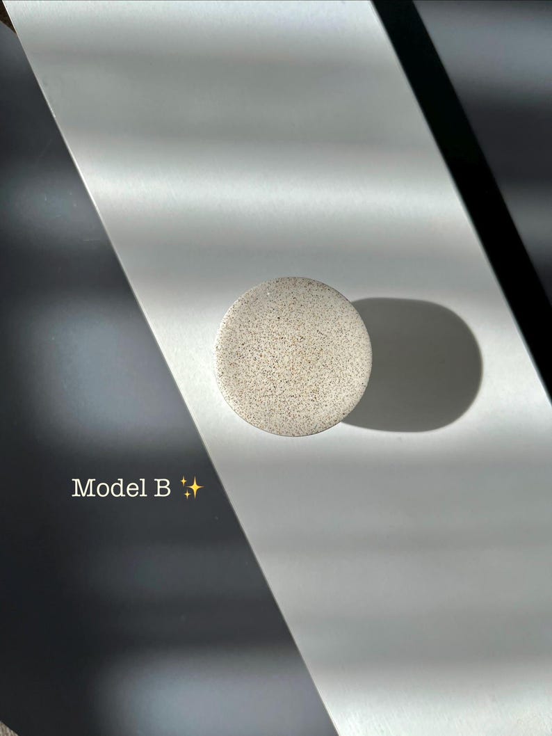 Puede incluir: Un objeto redondo, moteado, gris claro, etiquetado como "Modelo B" con un gr&aacute;fico de brillo. El objeto proyecta una sombra sobre una superficie blanca y gris. La imagen est&aacute; bien iluminada con sombras fuertes.