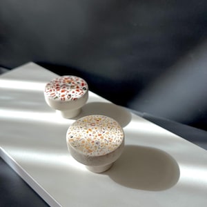Könnte beinhalten: Zwei dekorative Schrankknöpfe mit Terrazzo-Design. Die Knöpfe haben eine runde Oberseite mit einem gesprenkelten Muster aus Rot, Weiß und Beige. Der Sockel ist einfarbig hellgrau. Die Knöpfe befinden sich auf einer weißen Oberfläche mit schwarzem Hintergrund.