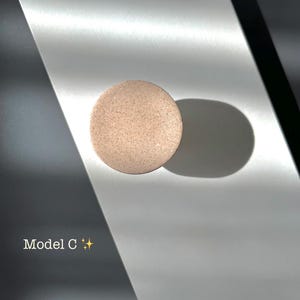 Puede incluir: Un objeto redondo y moteado, de color marr&oacute;n claro, posiblemente un producto cosm&eacute;tico, se encuentra sobre una superficie reflectante. El texto "Model C" es visible, junto con un peque&ntilde;o s&iacute;mbolo de estrella. La iluminaci&oacute;n crea sombras.