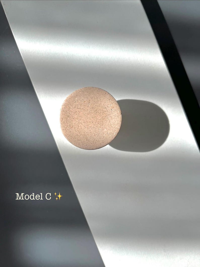 Puede incluir: Un objeto redondo y moteado, de color marr&oacute;n claro, posiblemente un producto cosm&eacute;tico, se encuentra sobre una superficie reflectante. El texto "Model C" es visible, junto con un peque&ntilde;o s&iacute;mbolo de estrella. La iluminaci&oacute;n crea sombras.