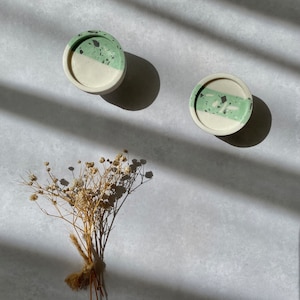 May include: Two small, round, green and white speckled concrete coasters. The coasters have a white base and a green top with black speckles. The coasters are on a gray surface with a bouquet of dried white flowers.