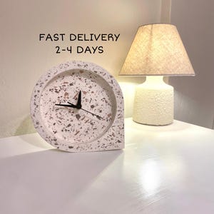 Könnte beinhalten: Weiße Terrazzo-Uhr mit schwarzen Zeigern. Die Uhr ist tropfenförmig und hat einen weißen Hintergrund mit braunen und schwarzen Flecken. Die Uhr steht auf einer weißen Oberfläche neben einer weißen Lampe mit einem beigefarbenen Lampenschirm. Der Text "FAST DELIVERY 2-4 DAYS" befindet sich über der Uhr.