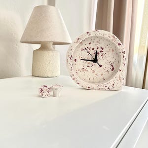 May include: A white terrazzo clock with black hands and a matching lamp and drawer pulls. The clock and drawer pulls have a speckled design with shades of pink and burgundy. The lamp has a textured, off-white base and shade.