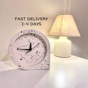 Könnte beinhalten: Weiße Terrazzo-Uhr mit schwarzen Zeigern. Die Uhr hat die Form einer Träne und ein minimalistisches Design. SCHNELLER VERSAND 2-4 TAGE steht über der Uhr.