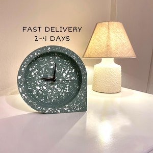 Könnte beinhalten: Eine grüne und weiße Terrazzo-Uhr mit schwarzen Zeigern. Die Uhr ist halbkreisförmig und hat einen weißen Sockel. Die Uhr steht auf einer weißen Oberfläche neben einer Lampe mit einem beigefarbenen Lampenschirm.