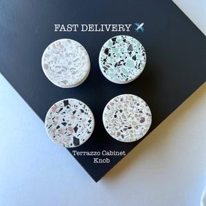 May include: Four terrazzo cabinet knobs in a variety of colors, including white, black, pink, and green. The knobs are round with a flat top and a white base. The text "FAST DELIVERY" and "Terrazzo Cabinet Knob" are visible.
