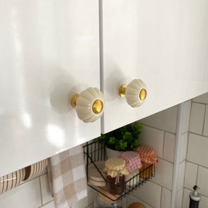 May include: Two white ceramic cabinet knobs with gold accents. The knobs are mounted on white cabinet doors.