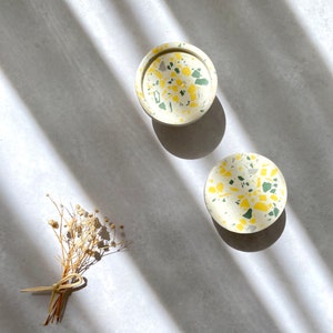 Może przedstawiać: Dwa małe okrągłe podkładki pod kubki z białej ceramiki z wzorem terrazzo w kolorze żółtym i zielonym.