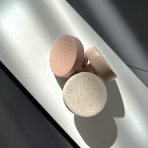 Puede incluir: Cuatro esponjas cosm&eacute;ticas redondas en tonos rosa y beige. Las esponjas est&aacute;n dispuestas sobre una superficie blanca, con sombras que crean contraste. Las esponjas parecen estar hechas de un material suave y poroso.