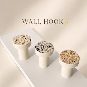 May include: Three cylindrical wall hooks with flat circular tops featuring a multi-colored terrazzo pattern of small stone fragments in shades of brown, purple, and green. The base of each hook is a solid matte off-white color. Text reads WALL HOOK.