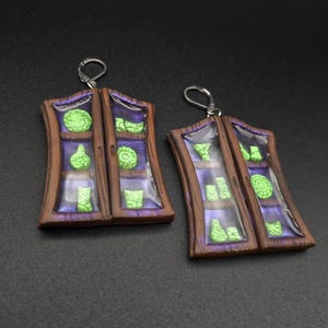 May include: A pair of dangle earrings shaped like miniature wooden cabinets with brown frames and purple interiors. Each cabinet features small, bright green sculpted objects arranged on shelves behind a clear, glossy resin surface.