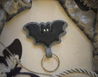 May include: A black bat-shaped keychain with a gray border and a silver keyring. The bat has white eye details. The keychain is displayed on a white surface with decorative elements, including a silver rope and butterfly cutouts.