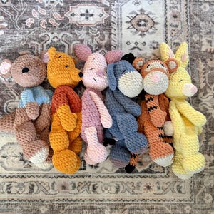 May include: A collection of handmade crochet stuffed animals, including a bear, kangaroo, pig, donkey, tiger, and rabbit. The toys are in various colors, such as yellow, orange, pink, and blue, and are arranged on a patterned rug.