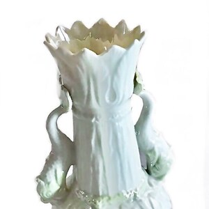 Op de afbeelding: Een witte keramische vaas met een geschulpte rand en een bolvormige voet met reliëfpatronen van schelpen. De vaas heeft twee decoratieve handvatten in de vorm van zwanenhalzen, met subtiele groene accenten op de zwanenfiguren en de voet.