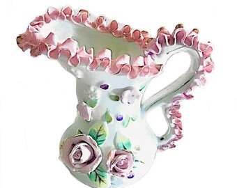May include: A white ceramic pitcher featuring a ruffled pink rim and handle. The body is decorated with three-dimensional sculpted pink roses, green leaves, and small painted floral accents. The pitcher has a curved spout and a glossy finish.