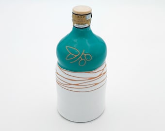 Può includere: Una bottiglia di olio d'oliva in ceramica con la parte superiore di colore verde turchese e la parte inferiore bianca. La bottiglia ha un disegno dorato di un ramo di ulivo con olive. L'etichetta sulla bottiglia recita "D.O.P. OLIO EXTRAVERGINE DI OLIVA CANINO".