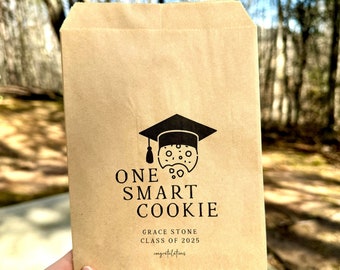 May include: A brown paper bag with a black graduation cap and cookie design. The text reads "One Smart Cookie", "Grace Stone", "Class of 2025", and "Congratulations".