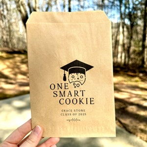 May include: A brown paper bag with a black graduation cap and cookie design. The text reads "One Smart Cookie", "Grace Stone", "Class of 2025", and "Congratulations".