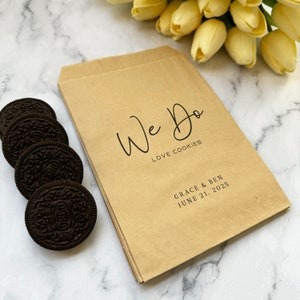 May include: Brown paper bags with the text "We Do Love Cookies" and the names "Grace & Ben" and the date "June 21, 2025".  The bags are stacked on top of each other.  There are four chocolate cookies in the lower left corner of the image.