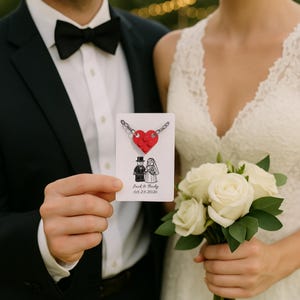 May include: A red heart-shaped pendant made of Lego bricks hangs from a silver chain. The pendant is on a white card with a drawing of a bride and groom and the date 05/23/2026. The card is held by a person wearing a black suit.