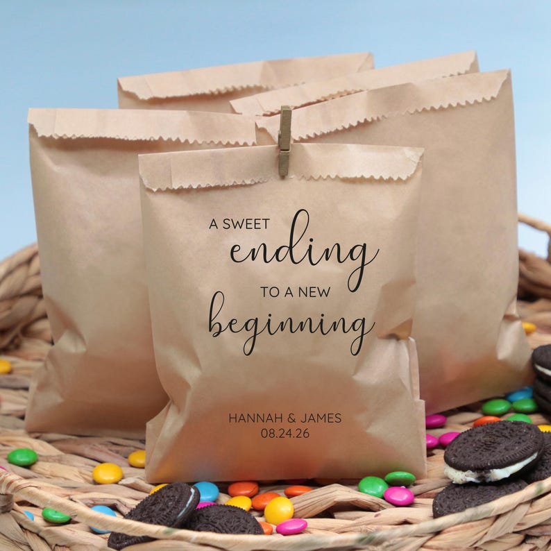 Op de afbeelding: Bruine papieren zakken met de tekst "A sweet ending to a new beginning" en de namen "Hannah & James" en de datum "08.24.26". De zakken zijn gevuld met kleine snoepjes en koekjes.