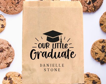 May include: A brown paper gift bag with a black graduation cap and the text "Our Little Graduate" with the name "Danielle Stone" printed below. The bag is surrounded by chocolate chip cookies.
