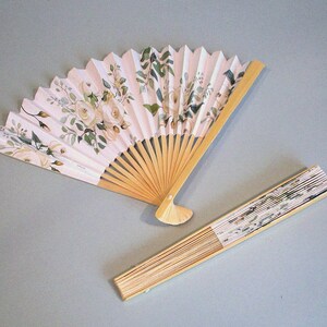 May include: Two hand fans with white paper and floral designs. The larger fan is open and has a wooden handle. The smaller fan is closed and has a wooden frame.