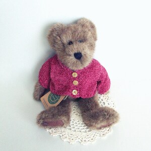 May include: A brown teddy bear wearing a magenta pink cardigan with three gold buttons. The bear is sitting on a cream-coloured, crocheted doily. A tag is attached to the bear.