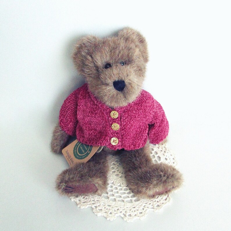 May include: A brown teddy bear wearing a magenta pink cardigan with three gold buttons. The bear is sitting on a cream-coloured, crocheted doily. A tag is attached to the bear.