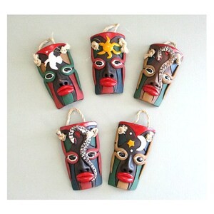 May include: Five small ceramic masks with different designs. Each mask is painted in vibrant colors, including red, green, black, white, and yellow. The masks feature various symbols, such as a sun, a moon, a star, and a snake.