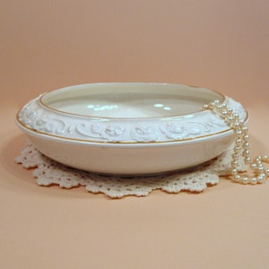 May include: White ceramic bowl with a gold rim and raised floral design. The bowl is sitting on a white crocheted doily. A strand of white pearls is draped over the edge of the bowl.