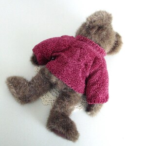 May include: A brown teddy bear wearing a magenta knitted sweater. The bear is lying on a white surface with a cream lace doily. The bear's fur is a mix of brown and tan.