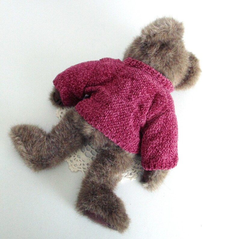 May include: A brown teddy bear wearing a magenta knitted sweater. The bear is lying on a white surface with a cream lace doily. The bear's fur is a mix of brown and tan.