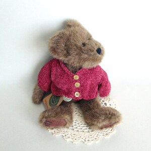 May include: A brown teddy bear wearing a magenta-coloured sweater with three gold buttons. The bear is sitting on a cream-coloured, crocheted doily. The bear has a black nose and a tag attached to its body.