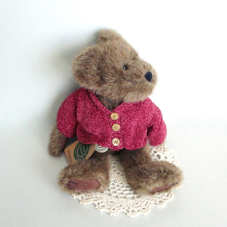 May include: A brown teddy bear wearing a magenta-coloured sweater with three gold buttons. The bear is sitting on a cream-coloured, crocheted doily. The bear has a black nose and a tag attached to its body.