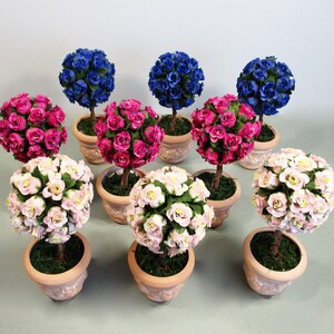 May include: Nine miniature potted topiary trees with pink, blue, and white paper roses. The trees are in terracotta pots with moss around the base.