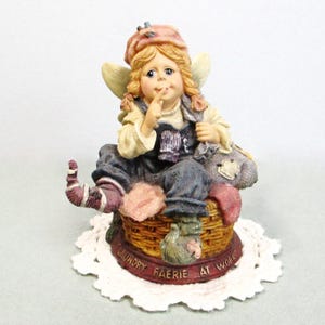 May include: A figurine of a fairy with blonde hair wearing a pink hat and blue overalls, sitting on a woven basket. The fairy holds a finger to their lips. The base is red with the text LAUNDRY FAERIE AT WORK embossed in gold lettering.