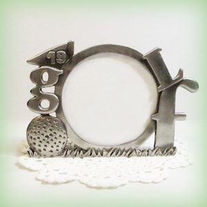 May include: A silver-toned picture frame shaped like a golf ball with the word "Golf" and the number "19" on it. The frame has a round opening for a photo.